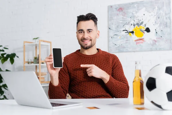Smiling gambler pointing at smartphone with blank screen near laptop, beer and blurred soccer ball — Stock Photo