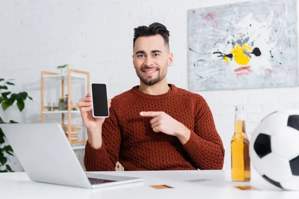 Giocatore d'azzardo sorridente che punta a smartphone con schermo vuoto vicino a laptop, birra e pallone da calcio offuscata — Foto stock