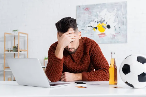 Corredor de apuestas deprimido sentado con la cabeza inclinada cerca de la computadora portátil, pelota de fútbol y botella de cerveza - foto de stock