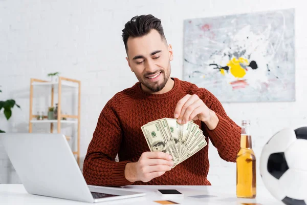 Happy bookmaker counting money near laptop, soccer ball and beer — Stock Photo