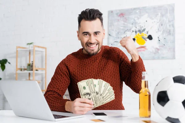 Excited bookmaker with dollar banknotes showing win gesture near laptop and soccer ball — Stock Photo