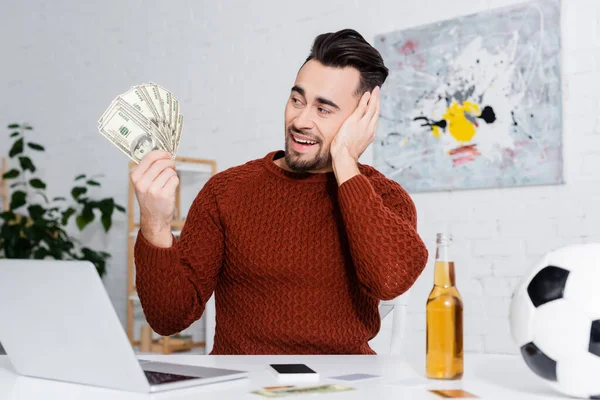 Amazed bookmaker holding dollars near laptop, bottle of beer and soccer ball — Photo de stock