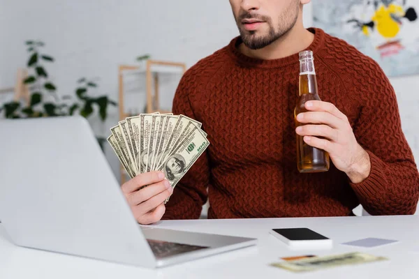 Partial view of gambler holding dollars and beer near blurred laptop — Stock Photo