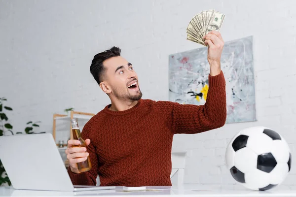 Astonished gambler holding dollars and bottle of beer near laptop and soccer ball — Photo de stock