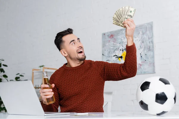 Astonished gambler holding dollars and bottle of beer near laptop and soccer ball — Stock Photo