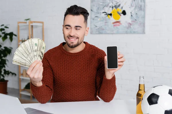 Joueur joyeux tenant dollars et smartphone avec écran blanc près du ballon de football — Photo de stock