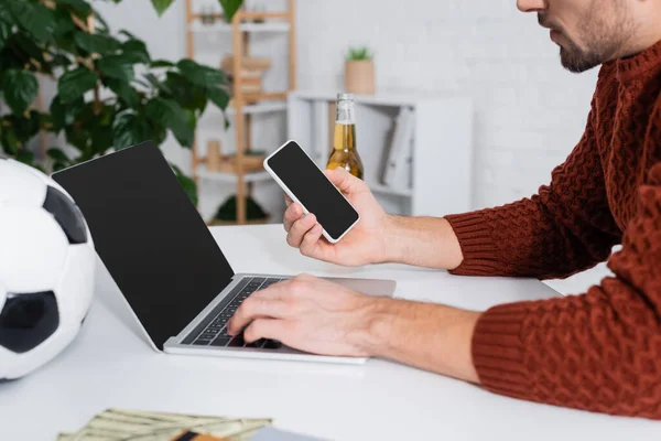 Cropped view of bookmaker with smartphone using laptop near blurred soccer ball — Stock Photo