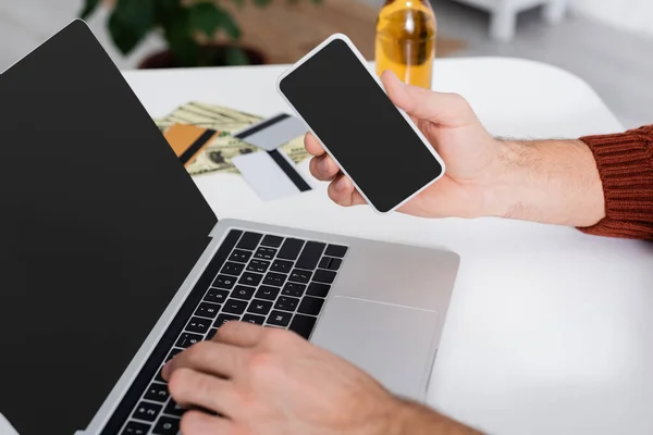 Partial view of bookmaker with smartphone and laptop with blank screen near blurred credit cards — Stock Photo