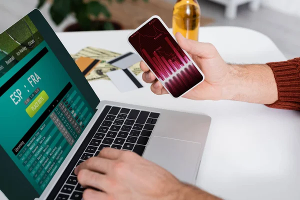 KYIV, UKRAINE - FEBRUARY 4, 2022: partial view of bookmaker holding smartphone with graphs near laptop with sportsbet website — Stockfoto