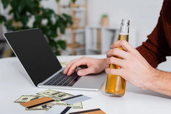 Vue recadrée du bookmaker avec bouteille de bière près d'un ordinateur portable, de l'argent et des cartes de crédit — Photo de stock