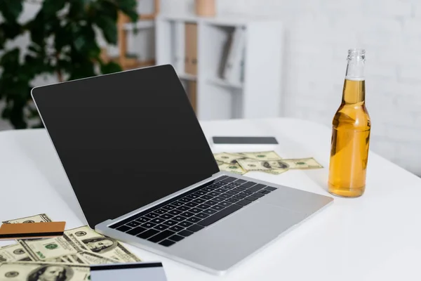 Portátil con pantalla en blanco cerca de tarjetas de crédito, dólares y botella de cerveza - foto de stock