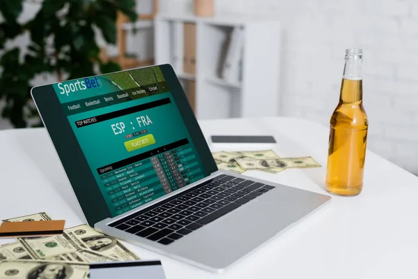 KYIV, UCRANIA - 4 DE FEBRERO DE 2022: tarjetas de crédito, billetes de dólar y cerveza cerca de la computadora portátil con sitio web sportsbet - foto de stock