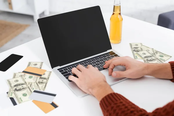 Vista parcial del jugador escribiendo en el ordenador portátil con pantalla en blanco cerca de dólares y tarjetas de crédito - foto de stock