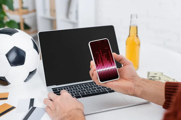 Partial view of bookmaker holding smartphone with analytics near laptop, soccer ball and credit cards - foto de stock