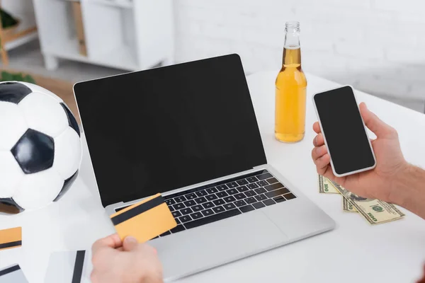 Vista parcial del jugador con tarjeta de crédito y teléfono inteligente cerca del ordenador portátil con pantalla en blanco - foto de stock