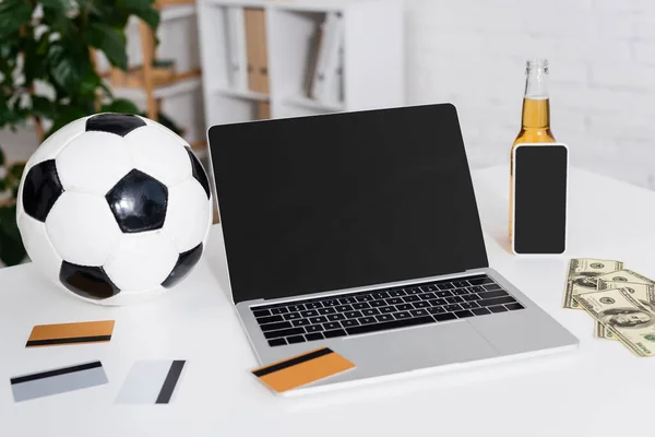 Portátil con pantalla en blanco cerca de pelota de fútbol, tarjetas de crédito, dólares y botella de cerveza - foto de stock