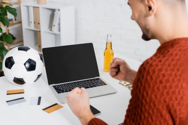 Blurred bookmaker holding clenched fists for luck near laptop, credit cards and soccer ball — Photo de stock