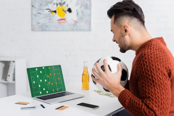 Side view of bookmaker with soccer ball near credit cards and laptop with sports game strategy on screen — Stock Photo