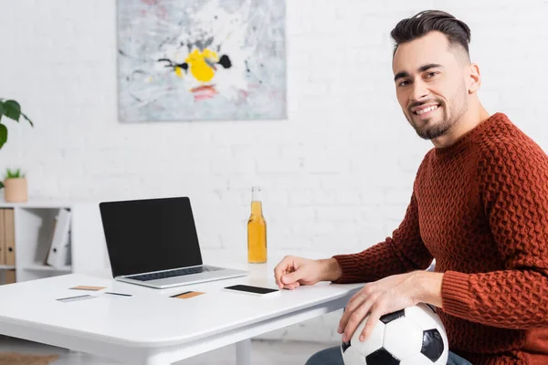 Jugador feliz con pelota de fútbol mirando a la cámara cerca de la computadora portátil y tarjetas de crédito - foto de stock