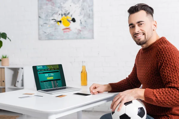 KYIV, UCRANIA - 4 DE FEBRERO DE 2022: corredor de apuestas feliz con pelota de fútbol mirando a la cámara cerca de la computadora portátil con el sitio web sportsbet - foto de stock