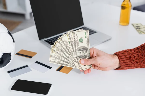 Cropped view of bookmaker holding dollars near laptop, credit cards and soccer ball — Stock Photo