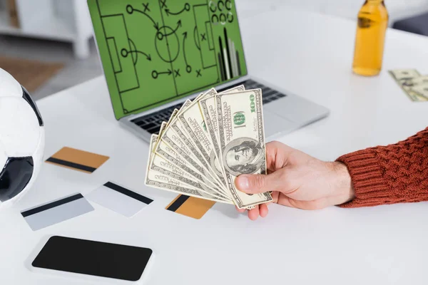 Cropped view of gambler with soccer ball near credit cards and laptop with sports game on screen — Stock Photo