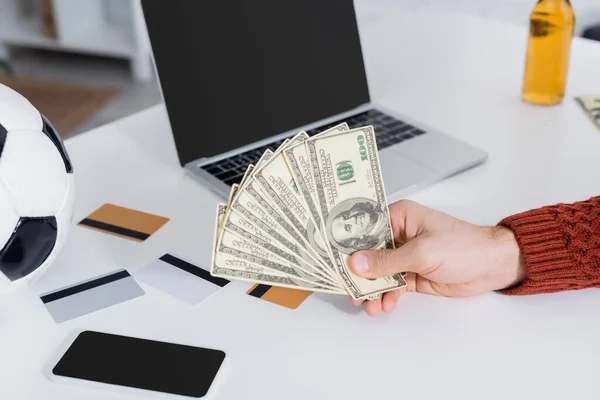 Partial view of bookmaker holding dollars near credit cards, soccer ball and laptop — Stock Photo