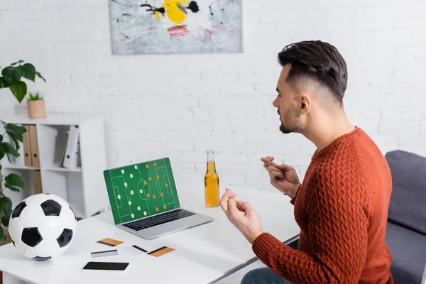 Vista laterale del giocatore d'azzardo con pallone da calcio vicino a carte di credito e laptop con strategia di gioco sportiva sullo schermo — Foto stock