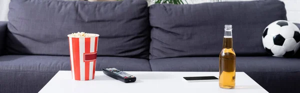 Canapé avec ballon de football près de la table avec seau de maïs soufflé, bière, télécommande tv et smartphone, bannière — Photo de stock