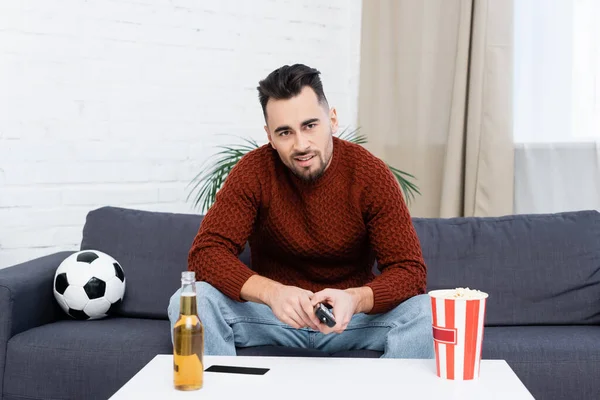 Skeptical sports fan watching championship on tv near soccer ball, popcorn and beer — Stock Photo