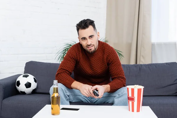 Skeptical sports fan watching championship on tv near soccer ball, popcorn and beer — Fotografia de Stock