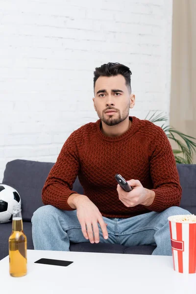 Sad sports fan with remote controller watching game on tv near soccer ball, beer and popcorn — Stock Photo