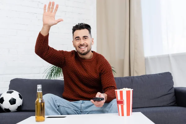 Cheerful sports fan waving hand while watching game at home near soccer ball and beer — Stock Photo