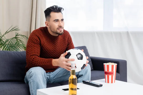 Concentrated man with soccer ball watching championship on tv near popcorn and beer — Photo de stock