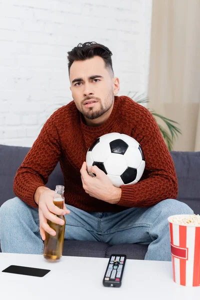 Fanático de los deportes tensos con pelota de fútbol y cerveza viendo campeonato en la televisión en casa - foto de stock