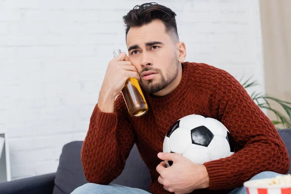 Molesto fanático de los deportes con pelota de fútbol y cerveza viendo partido en la televisión en casa - foto de stock