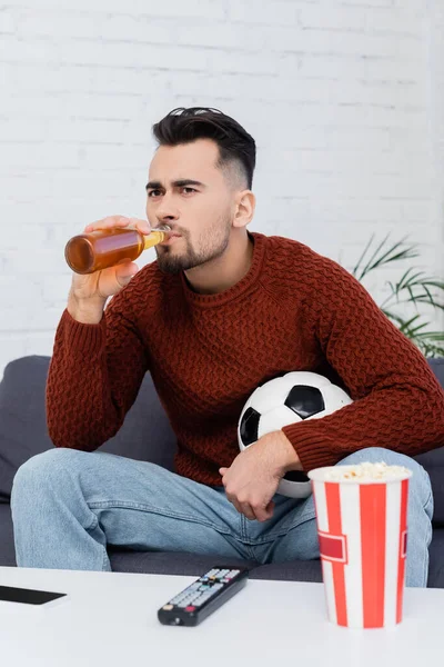 Fanático del fútbol enfocado beber cerveza mientras ve el juego en la televisión en casa - foto de stock