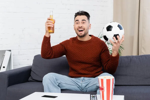 Fanático de los deportes emocionados con cerveza y pelota de fútbol viendo el juego en la televisión en casa - foto de stock