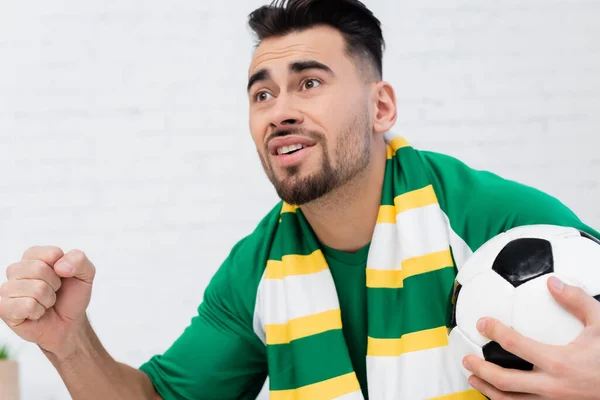 Excited sports fan holding soccer ball and clenched fist for luck while watching championship — Stock Photo