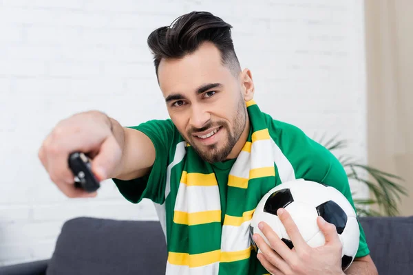 Smiling sports fan with soccer ball and blurred remote controller watching match on tv at home - foto de stock