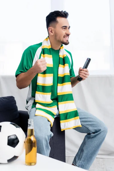 Hombre emocionado en bufanda rayada sosteniendo el puño apretado para la suerte mientras ve el partido de fútbol en casa - foto de stock