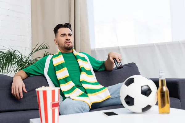 Sports fan in striped scarf watching game on tv near blurred soccer ball, beer and popcorn — Photo de stock