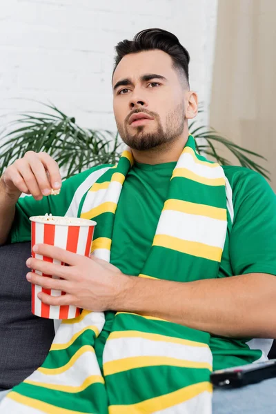 Focused man in striped scarf holding paper bucket of popcorn and watching sports game at home — Photo de stock