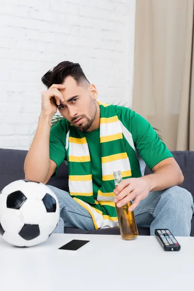 Frustrated sports fan with bottle of beer watching match on tv near soccer ball — Fotografia de Stock