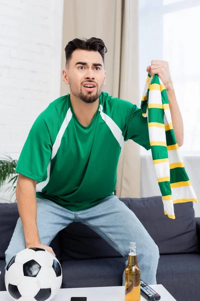 Preocupado y emocionado fanático de los deportes sosteniendo bufanda rayada mientras mira el partido de fútbol en la televisión - foto de stock