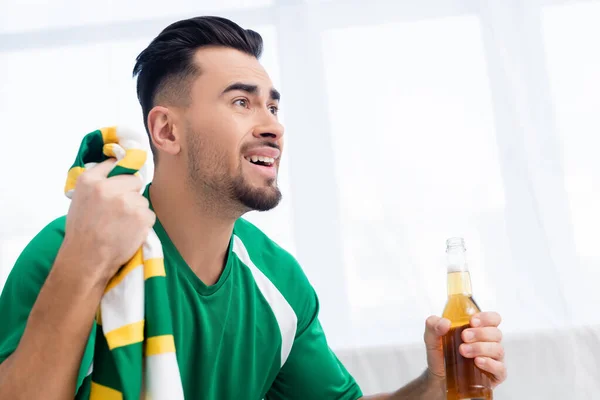 Worried sports fan holding striped scarf and bottle of beer while watching championship at home — Stock Photo