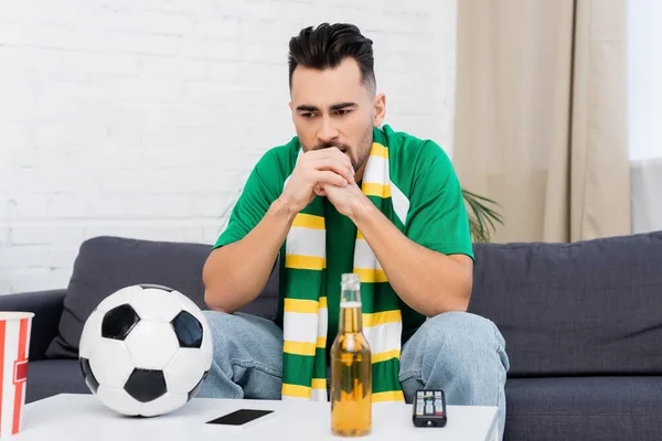 Frustrated sports fan sitting near tv remote controller, soccer ball and beer - foto de stock