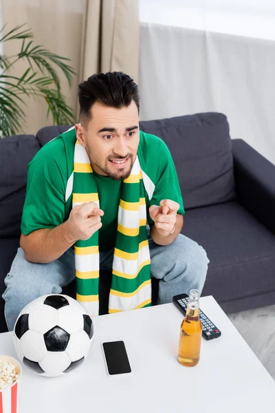 Homem tenso segurando dedos cruzados para a sorte perto de bola de futebol, cerveja e tv controle remoto — Fotografia de Stock