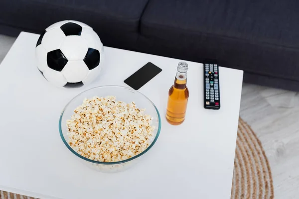 High angle view of bowl with popcorn near soccer ball, beer, tv remote controller and mobile phone on table — Stockfoto