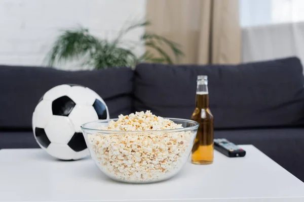 Tigela de pipoca perto de bola de futebol borrada, garrafa de cerveja e tv controle remoto na mesa — Fotografia de Stock